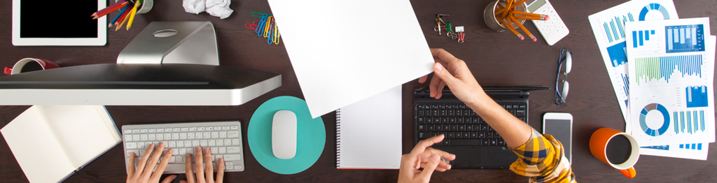 Overhead view of two workstations showing hands typing on keyboards, office supplies, and business analytics charts on dark desk