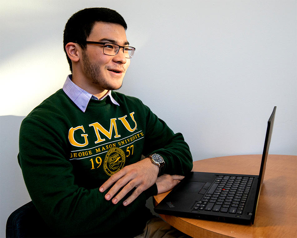 A Mason student with short dark hair wearing glasses and a green Mason sweatshirt over a collared button up shirt sits in front of a laptop and smiles while looking at the laptop
