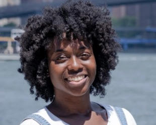 Julia Benedith wearing naturally curly hair and a denim jumper over a white t-shirt, smiles kindly at the camera.