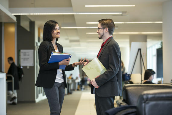 Two professionals conversing at a networking event, holding folders and documents, in a bright and modern office environment.