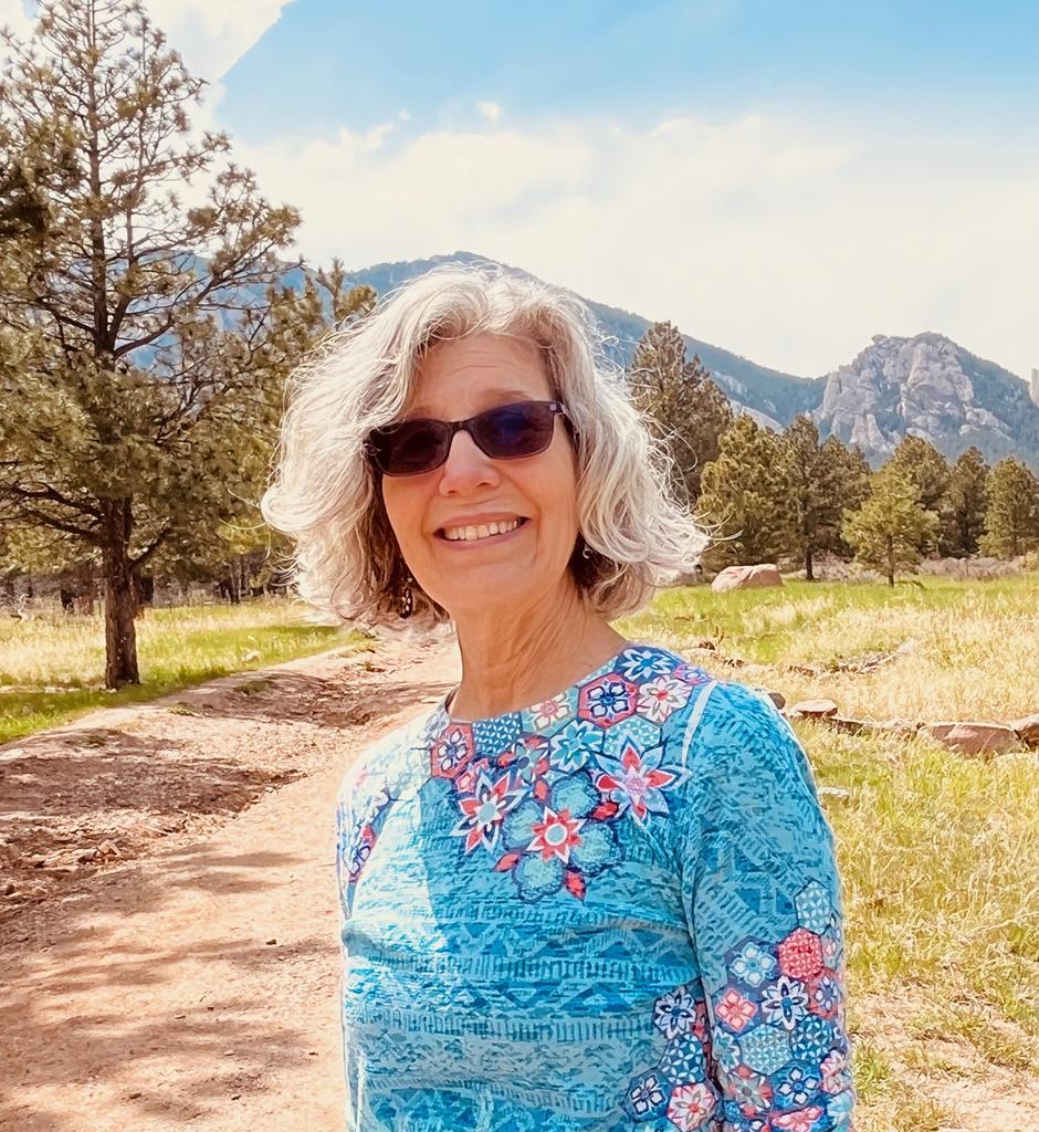Carolyn A. Wilson, Ph.D. stands on a trail in front of a mountain range, wearing a bright blue shirt.