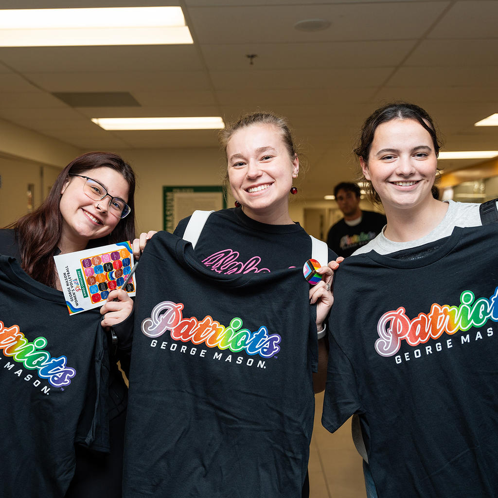 Students celebrate LGBTQ+ pride at George Mason University, smiling and holding rainbow Patriots shirts.