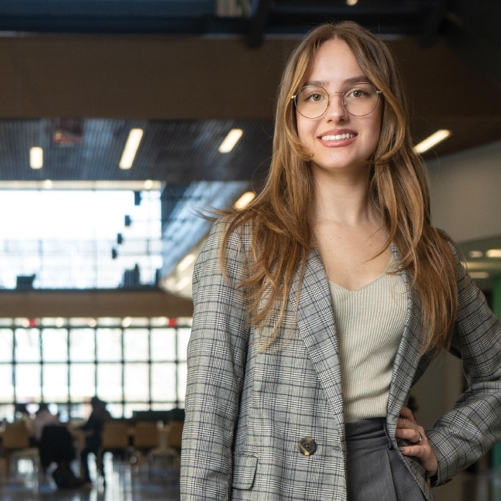 A George Mason student wearing a plaid blazer stands with one hand on their hip, smiling confidently at the camera